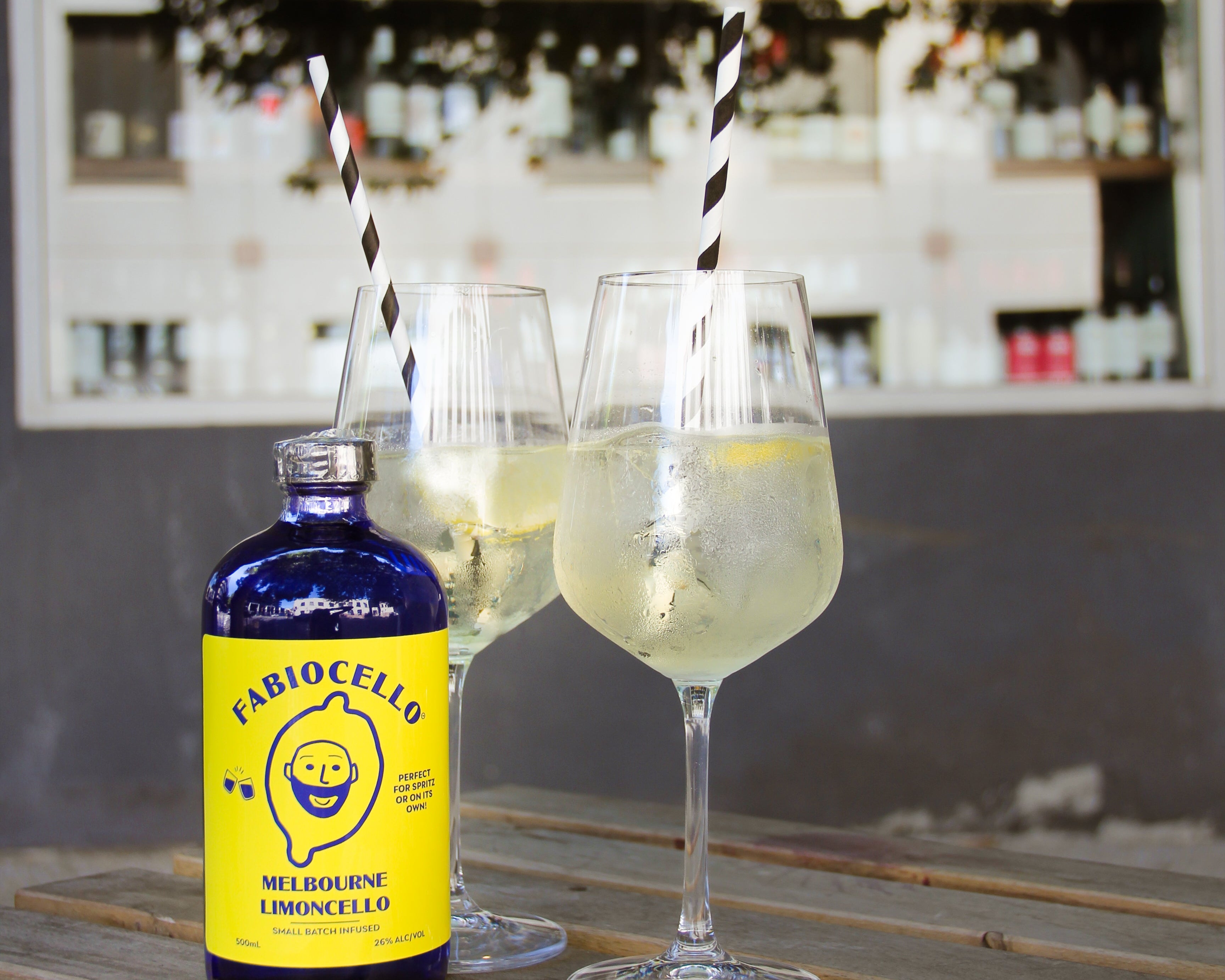 Bottle of Fabiocello limoncello with two glasses of limoncello on a wooden surface, bar setting in the background.